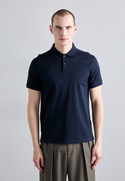 Polo shirt marine en coton doux, avec un col classique, une patte de boutonnage à deux boutons et un logo discret sur la poitrine. Associé à un pantalon ajusté.