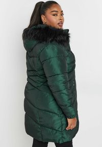 Grön puffjacka med en glänsande textur, utrustad med huva av svart fuskpäls, dragkedjefickor och diagonala quiltade sömmar för extra detalj.