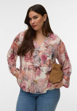 Kobieta z długimi ciemnymi włosami, ubrana w różową kwiecistą bluzkę z falbankami i niebieskie dżinsy, trzymająca w jednej ręce małą brązową zamszową torebkę.