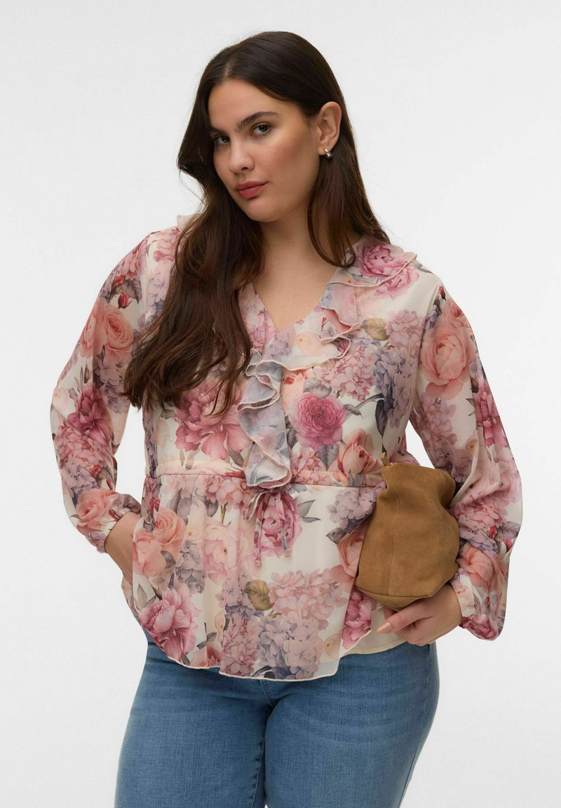 Femeie cu păr lung și închis la culoare, purtând o bluză roz cu volane și model floral și blugi albaștri, ținând o geantă mică maro din piele intoarsă într-o mână.