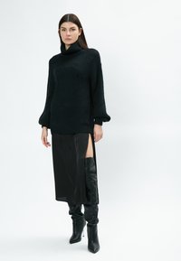 Schwarzer, übergroßer Strickpullover mit hohem Kragen, kombiniert mit einem schwarzen Satin-Midrock mit seitlichem Schlitz und knielangen Lederstiefeln.
