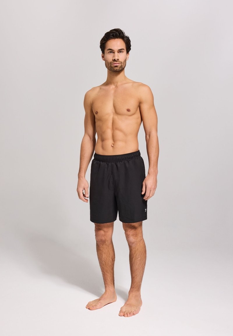 Schwarze Badeshorts mit elastischem Bund, knielangem Schnitt und logodetail an der Seite, getragen von einem oberkörperfreien Mann.