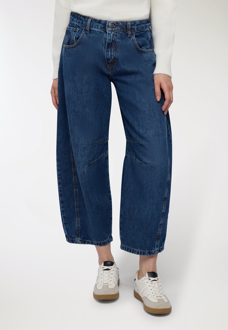 Jeans in denim blu a gamba larga con una finitura sbiadita, dotati di tasche frontali, cuciture a contrasto e design minimalista. Abbinati a sneakers bianche.