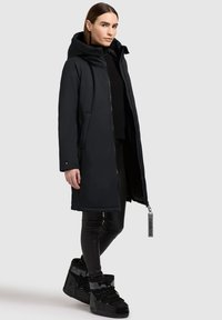 Zwarte waterdichte parka met een capuchon, ritsluiting, zijzakken en een gestructureerde binnenkant, gecombineerd met zwarte hoge laarzen.