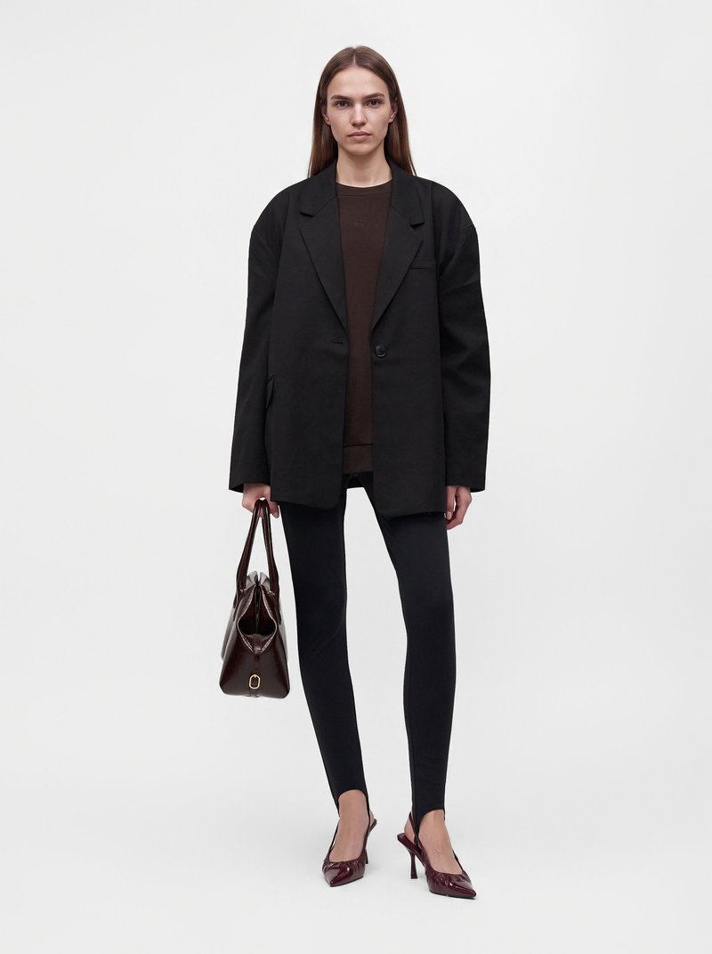 Femme portant un blazer noir oversize, des leggings noirs, des talons bordeaux, et portant un grand sac à main marron foncé, debout sur un fond blanc.