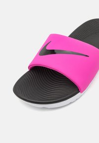 Chanclas Nike rosas con una tira ancha que presenta un logotipo negro. La plantilla texturizada negra tiene un patrón de olas, y una suela blanca acentúa el diseño.