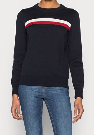 Femme portant un pull à manches longues noir avec des rayures horizontales blanches et rouges sur la poitrine et un jean bleu.