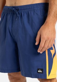 Șorturi de înot bleumarin cu talie elastică, șnur, și un design lateral galben strălucitor. Prezintă un logo negru pe piciorul stâng.