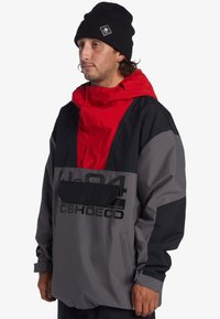 Šedý a čierny vonkajší pulóver s červenou kapucňou, ktorý má veľkú prednú kapsu a logá. Vyrobený z syntetického vodoodpudivého materiálu.