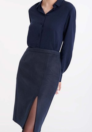 Frau trägt eine marineblaue Bluse mit Knöpfen und einen passenden marineblauen Bleistiftrock mit einem hohen Schlitz am Oberschenkel vor weißem Hintergrund.