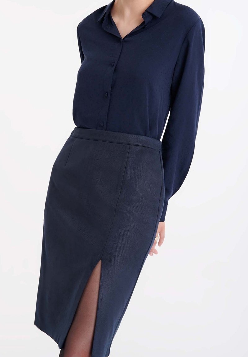 Femme portant un chemisier bleu marine à boutonnage et une jupe crayon bleu marine assortie avec une fente haute sur la cuisse, sur fond blanc.