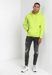 Sudadera con capucha de color amarillo neón, con un bolsillo delantero, combinada con pantalones skiny rasgados de color gris oscuro y zapatillas deportivas blancas.
