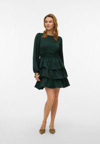 Tumši zaļa kleita ar garām piedurknēm, kārtainām volānām un jostasvietu ar gumiju. Gluda tekstūra, valkāta ar gaiši brūnām kurpēm ar smailiem purngaliem.