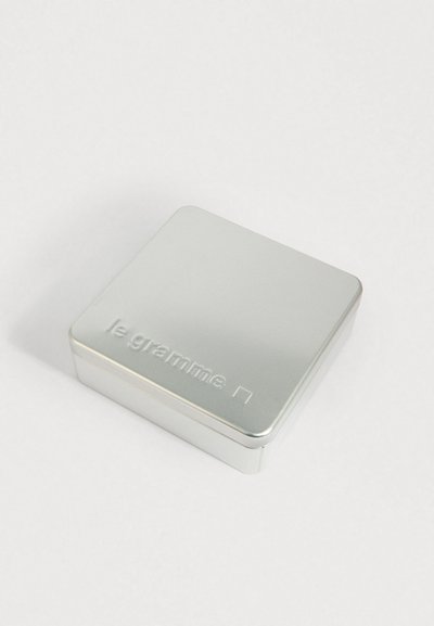 Väike hõbedane metallkast ümarate servadega, mille kaanel on reljeefne kiri "le gramme", asetatud valgele pinnale.