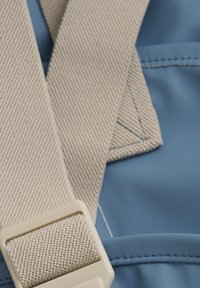 Beige vävd rem som korsar ett slätt blått material, fäst med en ljusfärgad plastspänne. Sytt detaljer är synliga.