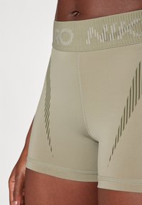 Pantaloni sportivi di colore verde oliva, con un elastico a coste in vita e logo bianco, materiale liscio e motivo a strisce diagonali.