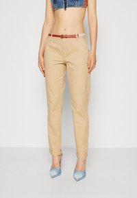 Pantalon chino beige avec revers, ceinturé d'une ceinture en cuir marron et top en denim court avec des accents orange et bleu. Escarpins bleus à bout pointu.