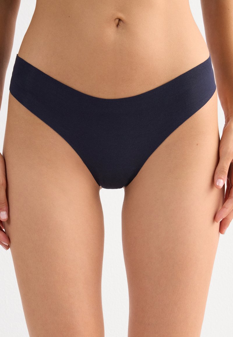 Femme portant un sous-vêtement bikini bas bleu foncé uni sans coutures, coupé au milieu du torse et sur le haut des cuisses, debout les mains détendues.