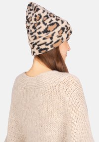 Bonnet en tricot à motif léopard dans des tons beige, noir et marron ; sommet pointu avec un revers roulé ; texture douce ; porté avec un pull en tricot beige clair.