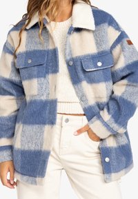 Frau in einer blauen und cremefarbigen karrierten Fleecejacke über einem weißen Strickpullover mit cremefarbenen hochgeschnittenen Hosen, die eine Hand in der Tasche hat.