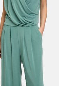 Combinaison avec col cheminé en tissu doux et texturé, de couleur teal, présentant des détails plissés et des poches latérales pour la fonctionnalité.