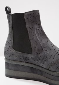 Botín de ante gris con suela de cuña, que presenta paneles laterales elásticos y detalles perforados. Textura suave y diseño elegante.