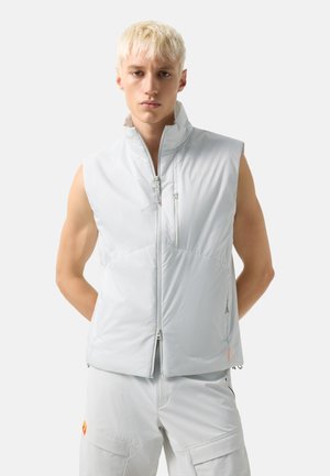 Junger Mann mit kurzen blonden Haaren, trägt eine ärmellose hellgraue Weste zum Reißverschluss und passende Cargo-Hosen, steht mit den Händen hinter dem Rücken.