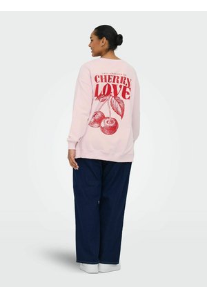 Vrouw gekleed in een roze sweatshirt met rode kersen en de tekst "Cherry Love" op de achterkant, gecombineerd met losse donkerblauwe broek en witte schoenen.