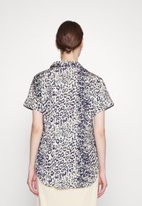 Blusa de mangas curtas em tecido leve, com um fundo creme e estampa de leopardo em roxo e preto, um colarinho e corte solto.