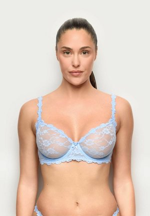 Donna con capelli castani raccolti, che indossa un reggiseno in pizzo azzurro chiaro, in piedi davanti a uno sfondo bianco semplice, rivolta verso l'obiettivo.