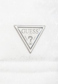 Fehér szövet dombornyomott mintával; ezüst háromszög alakú Guess logócímke, rajta a „WASHED” és „JEANS” feliratokkal, valamint egy kérdőjel szimbólummal.