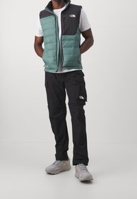 Padded grüne und schwarze Weste mit hohem Kragen, getragen über einem weißen T-Shirt, kombiniert mit schwarzen Cargo-Hosen und grauen Schuhen.