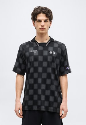 Νέος άνδρας που φοράει μαύρη καρό φανέλα ποδοσφαίρου Champion F.C. με λογότυπα στο στήθος και το μανίκι, στέκεται μπροστά σε απλό φόντο.