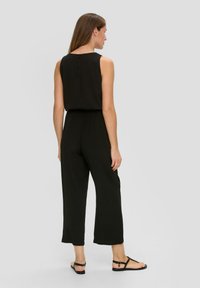 Zwarte jumpsuit met een mouwloos ontwerp, elastische tailleband en wijde broekspijpen. Gladde stof en knoopdetail op de rug.