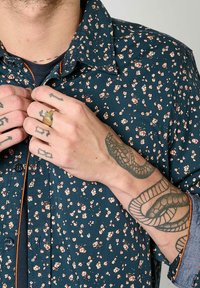 Donkergroene shirt met bloemenpatroon en oranje accenten en knopen. Voorzien van een knoopsluiting en opgerolde mouwen die tatoeages onthullen.