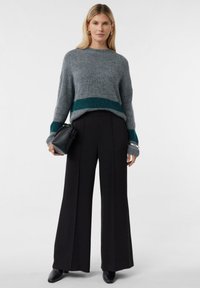 Kobieta w szarej wełnianej bluzie z teowymi paskami, czarnych szerokich spodniach, czarnych botkach, trzymająca małą czarną torebkę, na jednolitym tle.