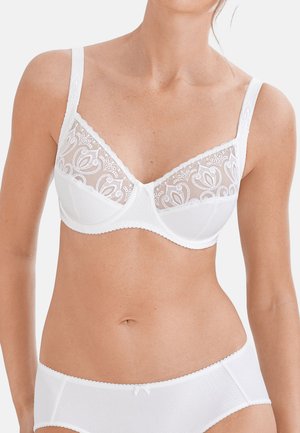 Kvinde iført hvid blonde-bøjle-bh med blomsterprint og matchende ensfarvede hvide trusser mod ensfarvet baggrund.