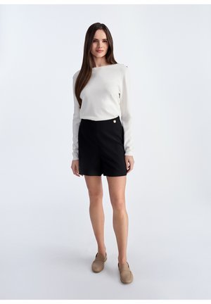 Biała bluzka z długim rękawem i guzikami, w połączeniu z czarnymi szortami o wysokim stanie. Modelka nosi brązowe buty typu slip-on, a jej włosy są proste.