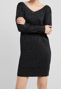 Abito in maglia nera con maniche lunghe, scollatura a V ampia e texture glitter. Design a lunghezza fianchi con spacchi laterali per facilitare i movimenti.