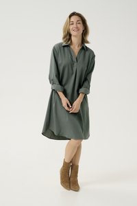 Groene, oversized jurk met een kraaglijn, lange opgerolde mouwen en een gebogen zoom. Draagt een paar bruine enkellaarsjes.