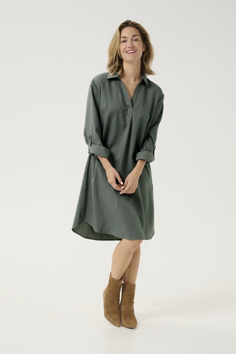 Groene, oversized jurk met een kraaglijn, lange opgerolde mouwen en een gebogen zoom. Draagt een paar bruine enkellaarsjes.