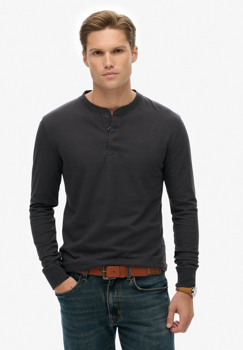 Ciemnoszara koszula typu henley z długim rękawem, z trzema brązowymi guzikami, okrągłym dekoltem i subtelnym logo. W połączeniu z niebieskimi dżinsami i brązowym paskiem.