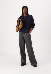 Navy gebreide pullover met een kraag en knoopsluiting, gecombineerd met grijze wijde jeans. Persoon met een glanzende bruine tas. Zwarte schoenen.