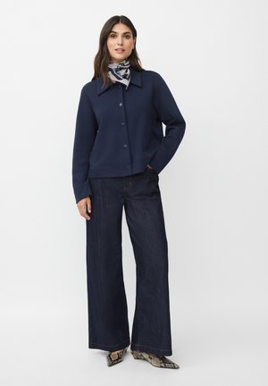 Veste cropped bleu marine avec un col et une fermeture à bouton, associée à un pantalon en denim foncé à jambes larges et des bottines à motifs.