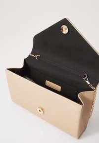 Bolso de imitación de piel beige con cierre de botón dorado y correa de cadena; el interior presenta forro negro y un bolsillo con una etiqueta de marca.