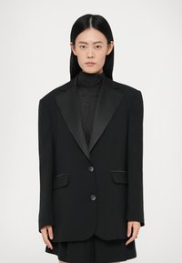 Zwarte blazer met satijnen pieklapels, twee knopen en zijzakken. Ontworpen met een rechte pasvorm en gestructureerde schouders. Soepele textuur.