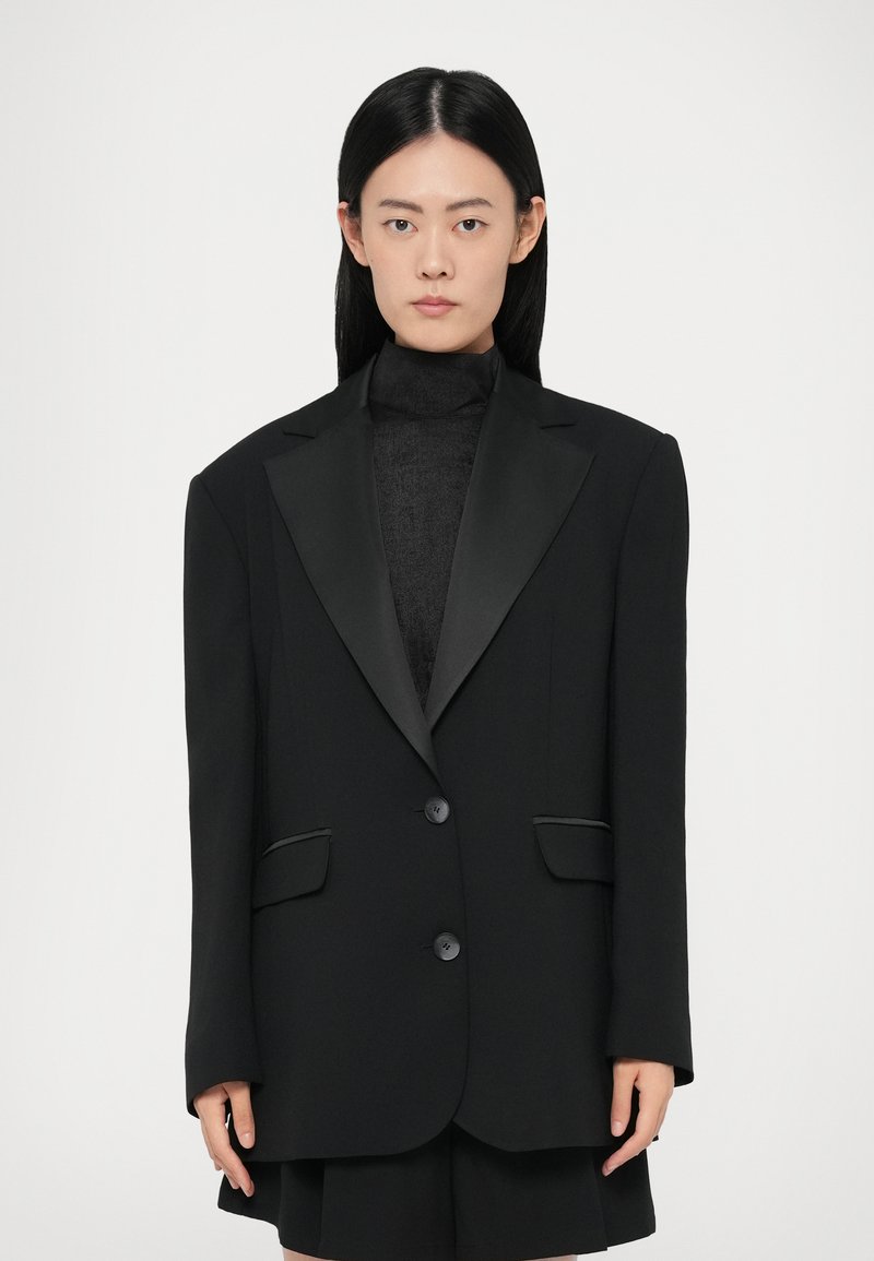 Zwarte blazer met satijnen pieklapels, twee knopen en zijzakken. Ontworpen met een rechte pasvorm en gestructureerde schouders. Soepele textuur.