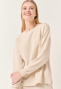 Maglione beige a maniche lunghe con una vestibilità ampia, collo rotondo, polsi a coste e spacchi laterali. Texture morbida e liscia con dettagli di cucitura minimi.