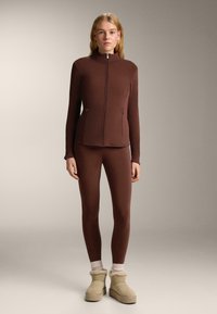 Smeđa sportskog kroja jakna s visokim ovratnikom i patentnim zatvaračem, u kombinaciji s odgovarajućim tajicama i bež gležnjačama s krznom. Mekana tekstura.