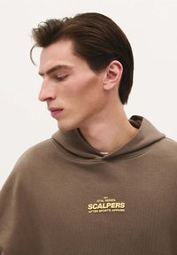 Brązowy hoodie z kapturem i miękką teksturą. Złote detale logo na klatce piersiowej przedstawiają "SCALPERS AFTER SPORTS APPAREL."